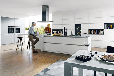 Expansive modern kitchen/diner in Surrey with flat-panel cabinets, white cabinets, stainless steel appliances, medium hardwood flooring and an island.