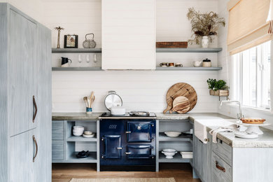 Photo of a mid-sized beach style u-shaped eat-in kitchen in Melbourne with a farmhouse sink, open cabinets, distressed cabinets, concrete benchtops, white splashback, shiplap splashback, coloured appliances, light hardwood floors, brown floor and grey benchtop.