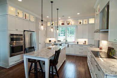 Design ideas for a mid-sized contemporary u-shaped open plan kitchen in Charlotte with a farmhouse sink, shaker cabinets, white cabinets, granite benchtops, white splashback, ceramic splashback, stainless steel appliances, medium hardwood floors, with island and brown floor.