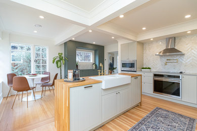 Example of a mid-sized transitional l-shaped medium tone wood floor, brown floor and coffered ceiling eat-in kitchen design in Other with a farmhouse sink, raised-panel cabinets, gray cabinets, wood countertops, beige backsplash, ceramic backsplash, stainless steel appliances, an island and brown countertops