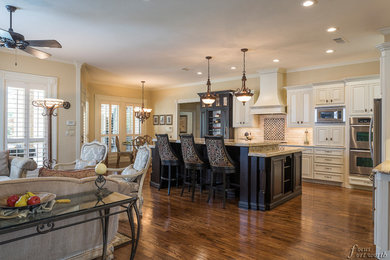 Example of a large classic galley medium tone wood floor open concept kitchen design in Dallas with a farmhouse sink, raised-panel cabinets, white cabinets, granite countertops, white backsplash, stone tile backsplash, stainless steel appliances and an island