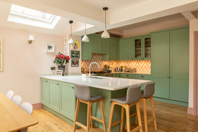 This is an example of a traditional u-shaped kitchen in Essex with a submerged sink, shaker cabinets, green cabinets, orange splashback, integrated appliances, medium hardwood flooring, a breakfast bar, brown floors and white worktops.