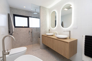Photo of a large contemporary master bathroom in Sydney with furniture-like cabinets, light wood cabinets, a freestanding tub, a curbless shower, a wall-mount toilet, white tile, ceramic tile, porcelain floors, a vessel sink, wood benchtops, grey floor, an open shower, a niche, a double vanity and a floating vanity.