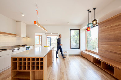 This is an example of a mid-sized contemporary galley eat-in kitchen in Melbourne with an integrated sink, flat-panel cabinets, light wood cabinets, stainless steel benchtops, white splashback, subway tile splashback, white appliances, light hardwood floors and with island.