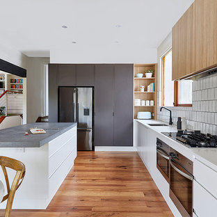 Photo of a large contemporary galley kitchen pantry in Melbourne with a drop-in sink, raised-panel cabinets, white cabinets, solid surface benchtops, white splashback, subway tile splashback, black appliances, plywood floors, with island and beige benchtop.