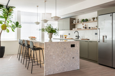 Inspiration for a contemporary galley kitchen in London with a belfast sink, flat-panel cabinets, green cabinets, white splashback, stainless steel appliances, light hardwood flooring, an island, beige floors and beige worktops.