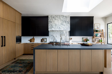 Eat-in kitchen - large contemporary l-shaped medium tone wood floor and brown floor eat-in kitchen idea in Los Angeles with a drop-in sink, flat-panel cabinets, black cabinets, soapstone countertops, white backsplash, marble backsplash, stainless steel appliances, an island and black countertops