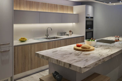 Contemporary single-wall kitchen/diner in Essex with flat-panel cabinets, light wood cabinets, glass sheet splashback, stainless steel appliances, laminate floors, an island and grey floors.