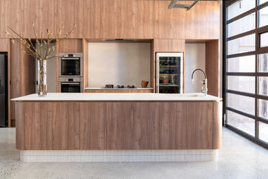 This is an example of a large modern galley open plan kitchen in Perth with an integrated sink, flat-panel cabinets, medium wood cabinets, solid surface benchtops, beige splashback, porcelain splashback, black appliances, concrete floors, multiple islands, grey floor and beige benchtop.