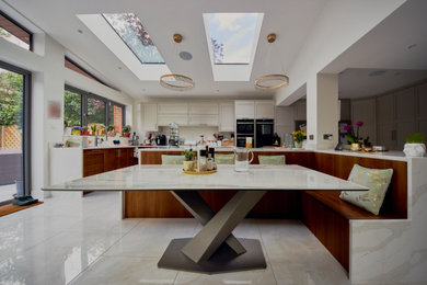 This is an example of a contemporary grey and cream kitchen in London with a submerged sink, medium wood cabinets, engineered stone countertops, beige splashback, black appliances, porcelain flooring, multiple islands, beige floors, grey worktops, a vaulted ceiling and feature lighting.