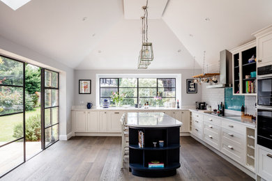 Traditional l-shaped open plan kitchen in London with a built-in sink, shaker cabinets, white cabinets, medium hardwood flooring, an island, white worktops, a vaulted ceiling and feature lighting.