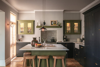 Inspiration for a transitional l-shaped open plan kitchen in London with a farmhouse sink, shaker cabinets, green cabinets, quartz benchtops, white splashback, stone slab splashback, medium hardwood floors, with island, brown floor and white benchtop.