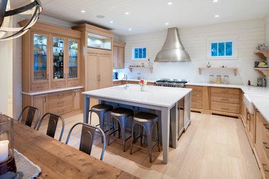 Large beach style u-shaped eat-in kitchen in Vancouver with a farmhouse sink, shaker cabinets, light wood cabinets, quartz benchtops, white splashback, stainless steel appliances, light hardwood floors, with island, beige floor and white benchtop.