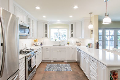 Photo of a mid-sized traditional u-shaped eat-in kitchen in Orange County with an undermount sink, raised-panel cabinets, white cabinets, quartz benchtops, white splashback, ceramic splashback, stainless steel appliances, porcelain floors and a peninsula.