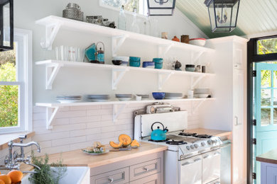 Photo of a small beach style l-shaped separate kitchen in Los Angeles with a farmhouse sink, grey cabinets, wood benchtops, white splashback, ceramic splashback, white appliances, medium hardwood floors, no island, brown floor, beige benchtop, recessed-panel cabinets and vaulted.
