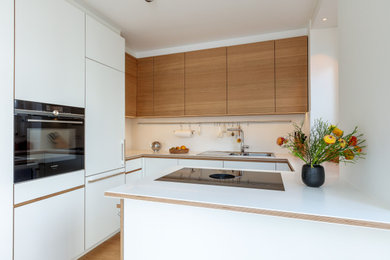 Photo of a mid-sized contemporary u-shaped open plan kitchen in Munich with a drop-in sink, flat-panel cabinets, white cabinets, solid surface benchtops, white splashback, black appliances, medium hardwood floors, with island, brown floor, white benchtop and recessed.