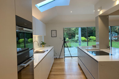 Contemporary open plan kitchen in Surrey with a double-bowl sink, flat-panel cabinets, grey cabinets, marble worktops, green splashback, glass sheet splashback, black appliances, light hardwood flooring, an island and white worktops.