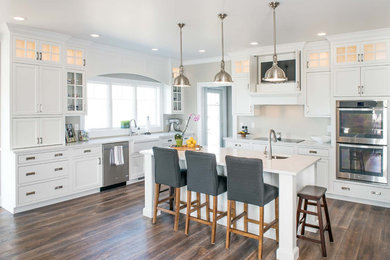 Design ideas for a mid-sized transitional u-shaped eat-in kitchen in Minneapolis with a farmhouse sink, shaker cabinets, white cabinets, quartz benchtops, stainless steel appliances, medium hardwood floors, with island and brown floor.