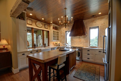 Large country u-shaped medium tone wood floor and brown floor open concept kitchen photo in Austin with a farmhouse sink, white cabinets, beige backsplash, stone slab backsplash, stainless steel appliances, an island, shaker cabinets and wood countertops