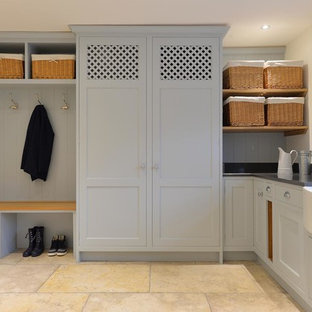 他の地域の大きいトラディショナルスタイルのおしゃれなランドリールーム (エプロンフロントシンク、ライムストーンの床、シェーカースタイル扉のキャビネット、グレーのキャビネット、白い壁、目隠し付き洗濯機・乾燥機、グレーのキッチンカウンター) の写真