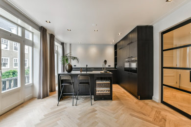 Photo of a medium sized modern cream and black l-shaped open plan kitchen in London with a built-in sink, flat-panel cabinets, black cabinets, marble worktops, black splashback, black appliances, light hardwood flooring, an island, brown floors, black worktops, a drop ceiling, marble splashback and a chimney breast.