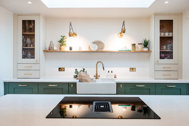 This is an example of a medium sized contemporary galley kitchen/diner in Surrey with a belfast sink, shaker cabinets, green cabinets, quartz worktops, white splashback, engineered quartz splashback, integrated appliances, laminate floors, an island, brown floors and white worktops.