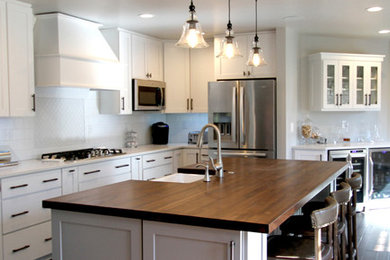 This is an example of a large contemporary single-wall eat-in kitchen in Kansas City with a farmhouse sink, raised-panel cabinets, white cabinets, wood benchtops, white splashback, ceramic splashback, stainless steel appliances, light hardwood floors, with island, grey floor and white benchtop.