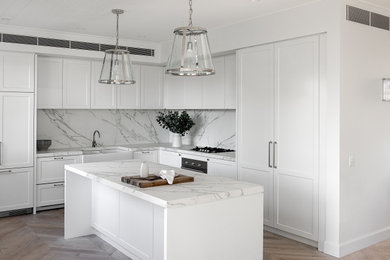 Photo of a mid-sized transitional l-shaped kitchen in Sydney with a farmhouse sink, shaker cabinets, white cabinets, white splashback, medium hardwood floors, with island, beige floor and white benchtop.
