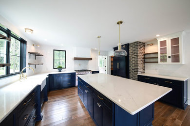 This is an example of a large country u-shaped kitchen pantry in Other with a farmhouse sink, blue cabinets, white splashback, medium hardwood floors, with island, white benchtop, shaker cabinets, quartz benchtops, subway tile splashback, panelled appliances and brown floor.