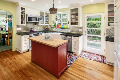 Kitchen - mid-sized eclectic l-shaped medium tone wood floor kitchen idea in Orange County with a farmhouse sink, stainless steel appliances, shaker cabinets, white cabinets, soapstone countertops, green backsplash, glass tile backsplash and an island