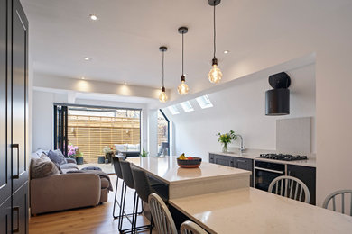 Design ideas for a small contemporary cream and black galley open plan kitchen in London with a built-in sink, recessed-panel cabinets, black cabinets, marble worktops, white splashback, marble splashback, black appliances, medium hardwood flooring, a breakfast bar, brown floors, white worktops and a chimney breast.