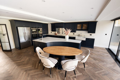 Photo of an expansive contemporary l-shaped open plan kitchen in Hertfordshire with a farmhouse sink, shaker cabinets, black cabinets, metallic splashback, glass sheet splashback, black appliances, medium hardwood floors, with island, brown floor and white benchtop.