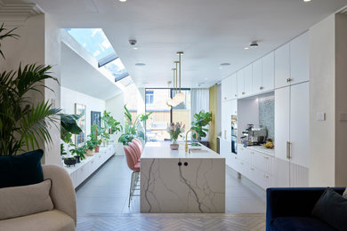 Example of a trendy ceramic tile and gray floor kitchen design in London with an undermount sink, flat-panel cabinets, white cabinets, solid surface countertops, travertine backsplash, white appliances, an island and white countertops