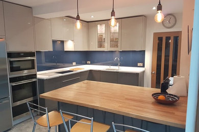 Medium sized contemporary l-shaped kitchen/diner in Kent with a built-in sink, flat-panel cabinets, grey cabinets, quartz worktops, blue splashback, glass sheet splashback, stainless steel appliances and an island.