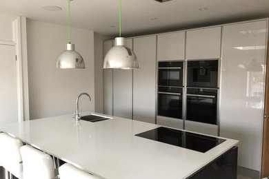 This is an example of a large modern kitchen in London with a built-in sink, flat-panel cabinets, grey cabinets, composite countertops, black appliances, ceramic flooring, an island and grey floors.