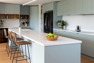This is an example of an expansive modern galley open plan kitchen in Melbourne with a drop-in sink, open cabinets, green cabinets, quartz benchtops, white splashback, stone tile splashback, black appliances, light hardwood floors, with island and white benchtop.
