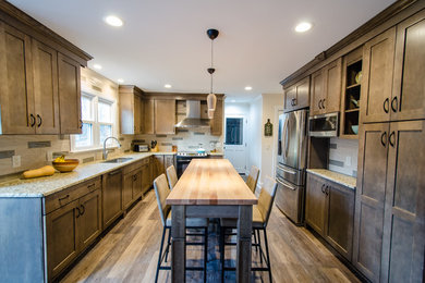 This is an example of a mid-sized arts and crafts galley open plan kitchen in Raleigh with an undermount sink, recessed-panel cabinets, brown cabinets, granite benchtops, beige splashback, stone slab splashback, stainless steel appliances, light hardwood floors and with island.
