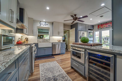 Photo of a large country l-shaped kitchen pantry in Miami with a farmhouse sink, shaker cabinets, blue cabinets, granite benchtops, beige splashback, shiplap splashback, stainless steel appliances, vinyl floors, a peninsula, brown floor and multi-coloured benchtop.