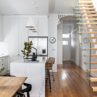 L-shaped eat-in kitchen in Sydney with an undermount sink, shaker cabinets, white cabinets, window splashback, stainless steel appliances, medium hardwood floors, with island, brown floor and white benchtop.