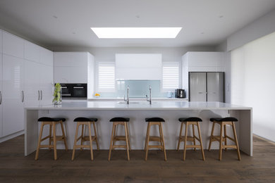 Expansive contemporary l-shaped open plan kitchen in Auckland with an integrated sink, flat-panel cabinets, white cabinets, solid surface benchtops, blue splashback, glass sheet splashback, stainless steel appliances, medium hardwood floors, with island, brown floor and white benchtop.