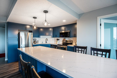 Example of a mid-sized eclectic galley vinyl floor and brown floor eat-in kitchen design in Edmonton with a double-bowl sink, flat-panel cabinets, blue cabinets, quartz countertops, white backsplash, porcelain backsplash, stainless steel appliances, an island and white countertops