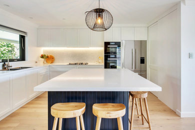 Design ideas for a large beach style l-shaped kitchen in Sydney with an undermount sink, shaker cabinets, white cabinets, quartz benchtops, mosaic tile splashback, black appliances, light hardwood floors and white benchtop.