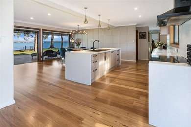 This is an example of a large contemporary single-wall eat-in kitchen in Sydney with a farmhouse sink, flat-panel cabinets, beige cabinets, granite benchtops, white splashback, black appliances, light hardwood floors, with island, brown floor and white benchtop.