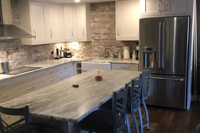 Eat-in kitchen - large farmhouse l-shaped medium tone wood floor and brown floor eat-in kitchen idea in Grand Rapids with a farmhouse sink, recessed-panel cabinets, white cabinets, granite countertops, gray backsplash, brick backsplash, stainless steel appliances and an island