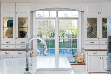 Large transitional single-wall light wood floor and brown floor open concept kitchen photo in Other with a farmhouse sink, raised-panel cabinets, white cabinets, quartz countertops, white backsplash, ceramic backsplash, stainless steel appliances, an island and white countertops