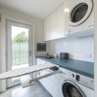 Photo of a small contemporary galley separated utility room in Canberra - Queanbeyan with a single-bowl sink, raised-panel cabinets, white cabinets, laminate countertops, white walls, concrete flooring, a stacked washer and dryer, grey floors and blue worktops.