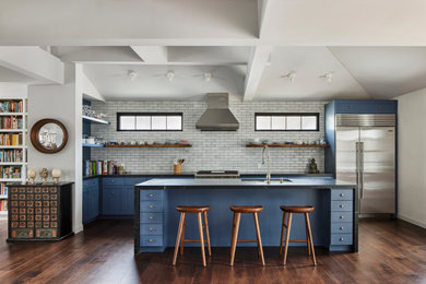 Photo of a mid-sized transitional u-shaped open plan kitchen in Los Angeles with a double-bowl sink, shaker cabinets, blue cabinets, soapstone benchtops, grey splashback, ceramic splashback, stainless steel appliances, medium hardwood floors, with island, brown floor, black benchtop and exposed beam.