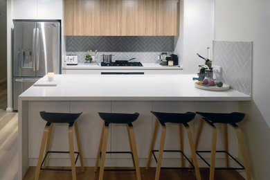 Photo of a mid-sized modern galley kitchen pantry in Sydney with an undermount sink, flat-panel cabinets, white cabinets, quartz benchtops, grey splashback, ceramic splashback, black appliances, light hardwood floors, a peninsula, brown floor and white benchtop.