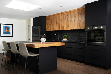 Inredning av ett modernt mellanstort svart svart kök, med en undermonterad diskho, släta luckor, svarta skåp, svart stänkskydd, stänkskydd i sten, svarta vitvaror, mellanmörkt trägolv, en köksö och brunt golv