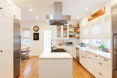 Example of a mid-sized trendy l-shaped light wood floor and beige floor eat-in kitchen design in New York with a farmhouse sink, recessed-panel cabinets, white cabinets, quartzite countertops, white backsplash, subway tile backsplash, white appliances and an island
